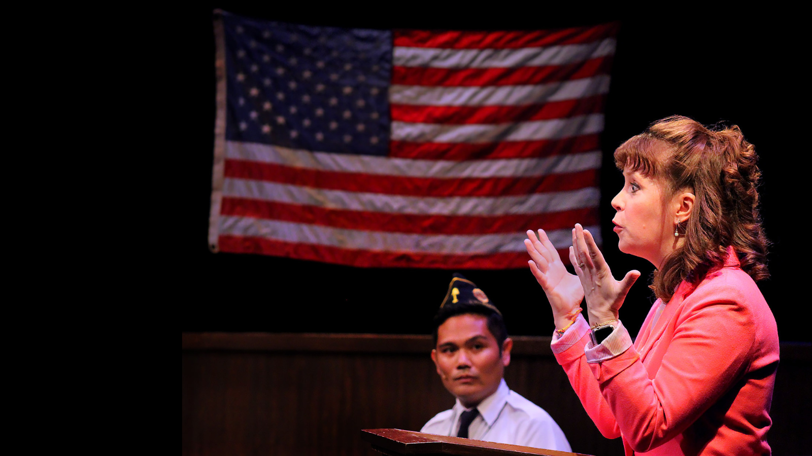 production photo of a woman at a podium with an American Legion male in the background underneath the American flag