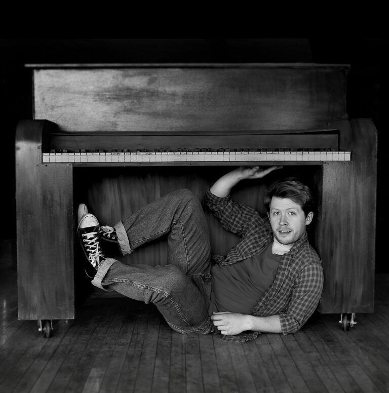 black and white photo of a young man crouched underneath an upright piano