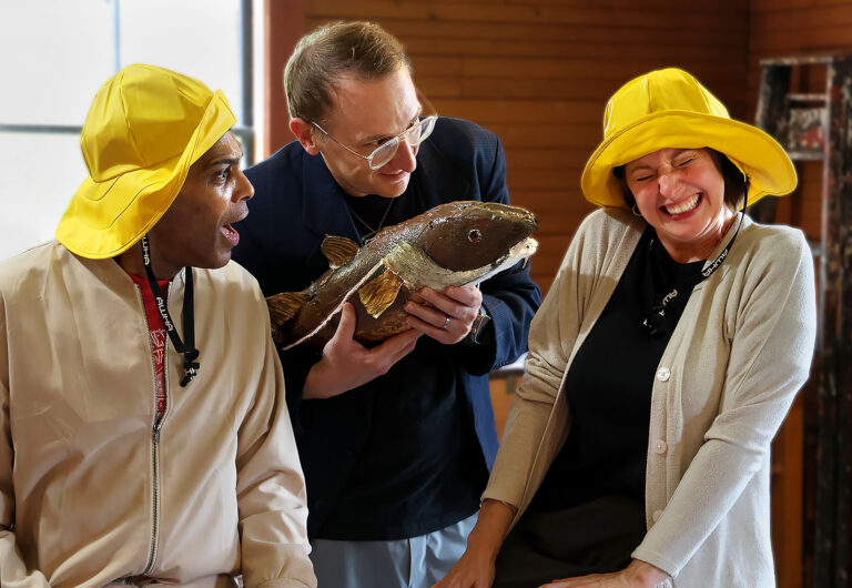 Man and woman seated in yellow hats being taunted by man standing with a cod fish