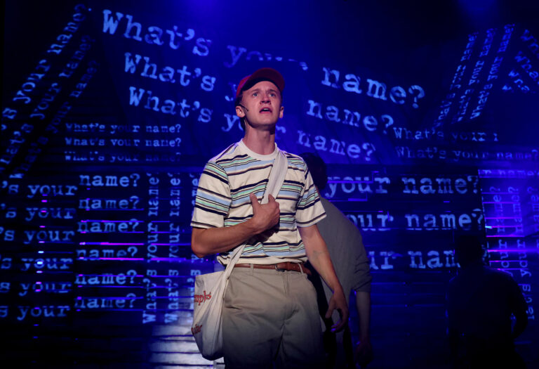 young man in baseball cap standing against projections reading "what's your name"